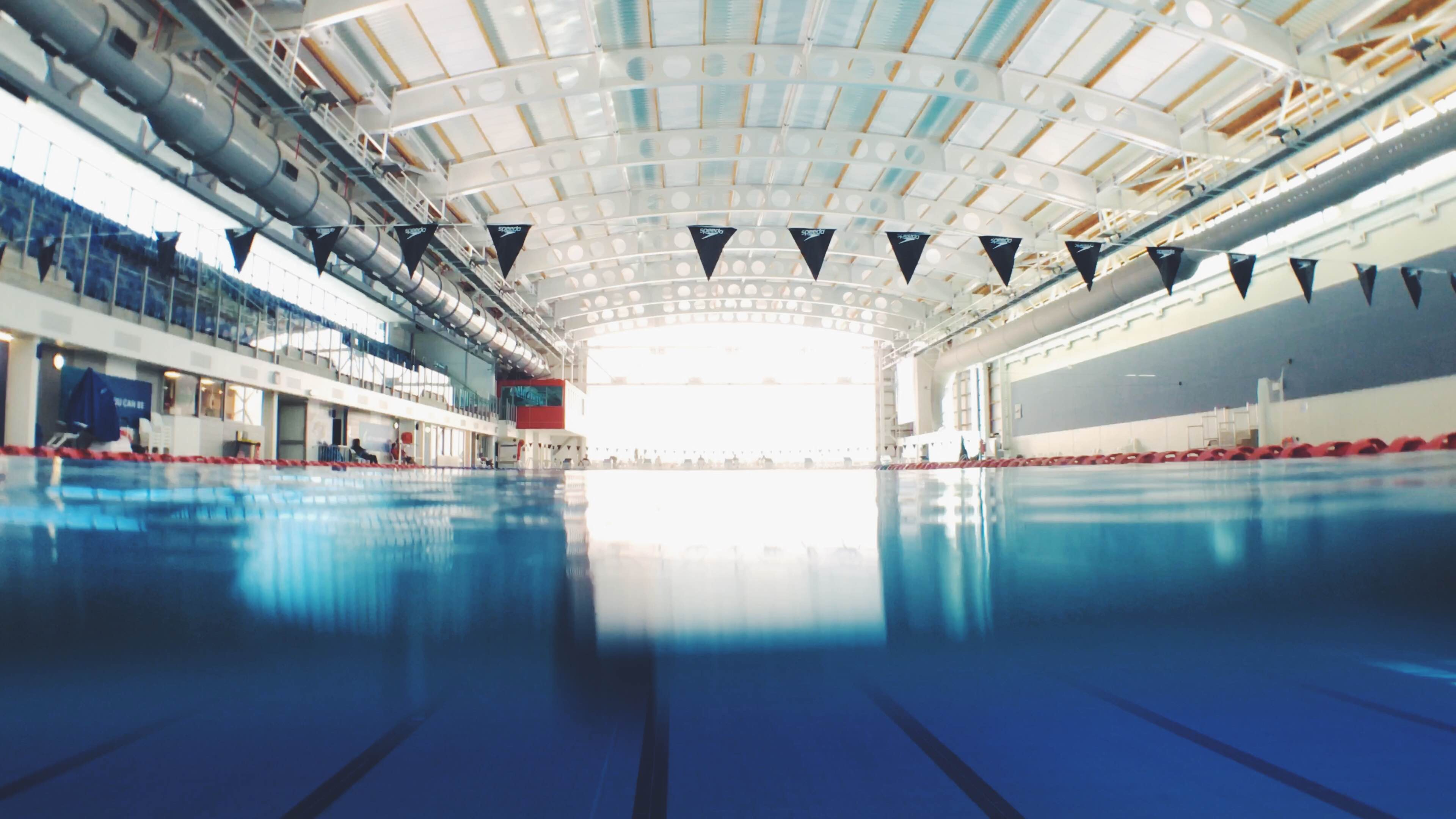 Image size 1600x900. Illustrative style. A calm indoor pool with a beginner swimmer practicing freestyle, soft morning light reflecting on the water, simple and encouraging training mood.