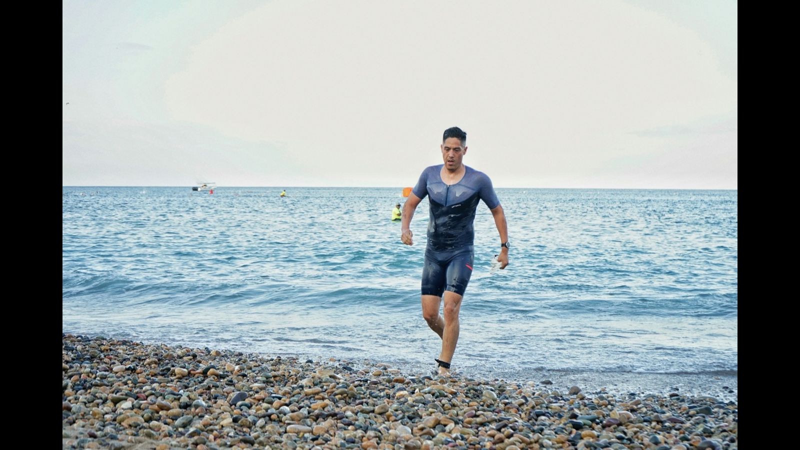 Image size 1600x900. Triathlete running out of open water toward a pebble beach during a race.