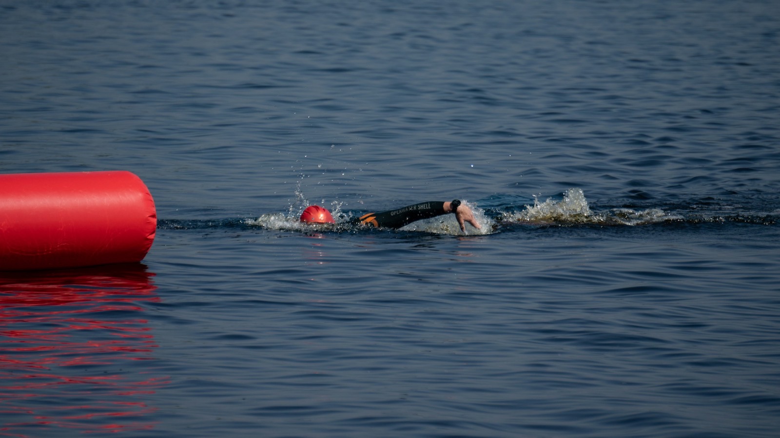 Image size 1600x900. Open-water swimmer in a wetsuit passing a red buoy on calm water.